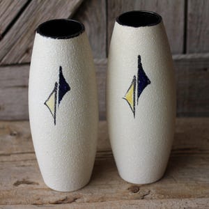 May include: Two white ceramic vases with a textured surface and black and yellow geometric designs. The vases are tall and slender.