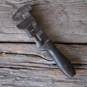 May include: A vintage adjustable wrench with a dark, weathered metal finish. The tool features a sliding jaw mechanism, a knurled adjustment nut, and a solid, rounded handle. The metal surface shows signs of age, oxidation, and a deep gray patina.