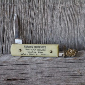 May include: A vintage folding pocket knife with a cream-colored handle featuring black text that reads CARLTON ANDERSON'S DEEP ROCK SERVICE Goodyear Tires AAA - Phone 817 - Willmar. The knife has a single silver blade and a brass horse head ornament.