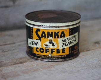 May include: Vintage Sanka coffee can with a black and yellow label. The label reads "Sanka New Coffee Improved Flavor" with an illustration of a person. The can is cylindrical with a dark brown lid.