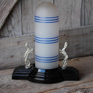 May include: An Art Deco-style lamp with a frosted white glass shade adorned with three horizontal blue stripes. The lamp sits on a black base with silver-colored deer figurines on each side. The design is reminiscent of the 1930s.