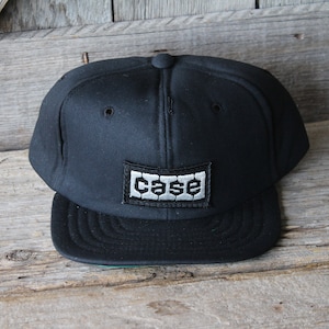 May include: Black baseball cap with a flat brim. The cap features a rectangular patch with the word "CASE" in a stylized font. The patch is white with a black border, and the cap is set against a wooden background.