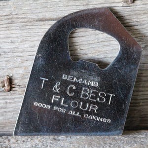 May include: A vintage metal flour scraper with a curved handle and a rectangular blade. The text "DEMAND T & C BEST FLOUR GOOD FOR ALL BAKINGS" is embossed on the blade. The scraper is set against a weathered wooden surface.