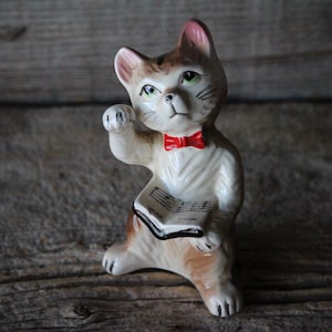 May include: A ceramic cat figurine with a red bow tie, holding a book of sheet music and raising a paw. The cat has brown and white fur, green eyes, and black whiskers. A decorative item.