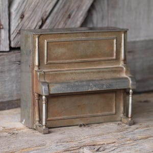May include: A small, antique-style piano figurine in shades of bronze and silver. The miniature piano features detailed molding and decorative legs, set against a rustic wooden backdrop. A charming decorative piece for music lovers or collectors.