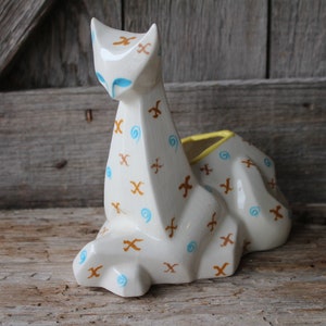 May include: White ceramic cat planter with brown and blue geometric patterns. The planter has a yellow rim and is shaped like a stylized cat.
