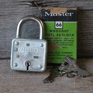 May include: A silver-colored Master 66 wrought steel padlock with a curved handle, keyhole, and two keys. The padlock is next to its original green and black box, which has the Master logo and product information.