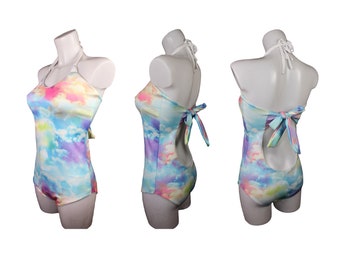Op de afbeelding: Een halter-badpak met een kleurrijke wolkendruk en een strik aan de achterkant. Het badpak is wit met roze, blauwe en gele wolken.
