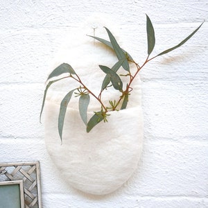 May include: A white felt wall pocket with a curved shape, holding eucalyptus branches with green leaves and small buds. The pocket is hanging on a white brick wall, creating a natural, minimalist aesthetic.