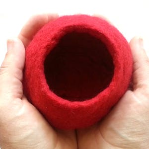 May include: A red felt bowl with a smooth, rounded interior. The bowl is held in a person's hands.