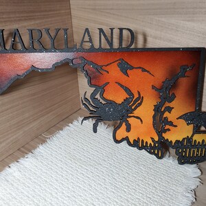 May include: A black metal cutout of the state of Maryland with a sunset gradient background. The state name is written above the outline. A black crab and a sailboat are cut out of the state shape.