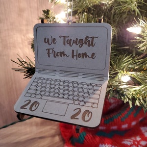 Pode incluir: Um enfeite de Natal de madeira em forma de computador portátil com o texto "We Taught From Home 2020" na tela.