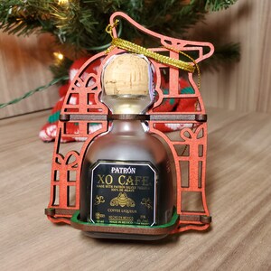May include: A red wooden Christmas ornament shaped like a house with a small bottle of Patron XO Cafe coffee liqueur inside. The bottle is labelled with the text "Patron XO Cafe Made with Patron Silver Tequila 100% de Agave Coffee Liqueur 35% alc/vol. Hecho en Mexico Made in Mexico."