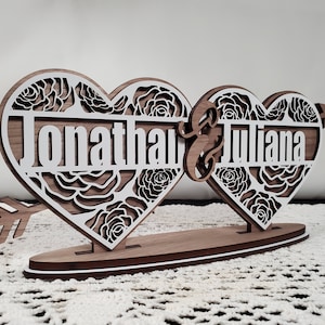 May include: Wooden wedding centrepiece featuring two interlocking hearts with a floral design and the names "Jonathan & Juliana" inscribed on them. The hearts are attached to a wooden base with an arrow.