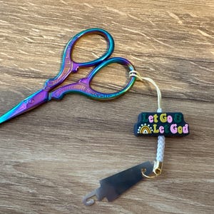 Puede incluir: Tijeras de bordar iridiscentes con acabado arcoíris, junto a un pequeño dije que dice "Let Go & Let God". Las tijeras tienen un mango decorativo y una hoja afilada y puntiaguda. Un pequeño cortahilos está adjunto.