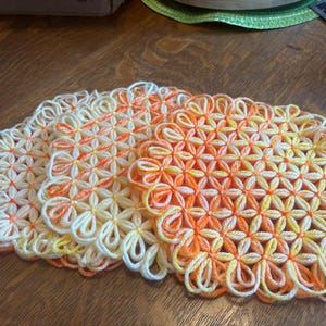 Puede incluir: Tres tapetes de ganchillo redondos hechos a mano con un patrón repetitivo en forma de flor. El hilo cambia de color de blanco a amarillo y naranja, creando un efecto de degradado vibrante en los bucles interconectados de cada pieza.
