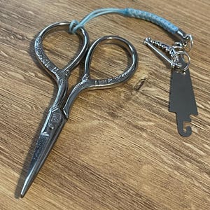 Puede incluir: Tijeras de bordar plateadas con mangos ornamentados y una pequeña etiqueta a juego. Las tijeras tienen un cordón trenzado azul y blanco adjunto. Las tijeras están sobre una superficie de madera.