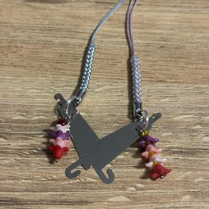 May include: A gray metal charm with two umbrella shapes, each with a beaded flower charm in shades of pink, purple, and red. The charm has two braided silver and lavender cords for attachment.