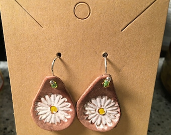 May include: A pair of brown leather earrings with a white daisy design and green beads. The earrings are hanging on a brown card.
