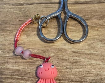 Könnte beinhalten: Silberne Stickschere mit einem rosa-roten Perlenanhänger. Der Anhänger zeigt eine lächelnde rosa Qualle mit Kulleraugen. Die Schere ist geöffnet und liegt auf einer Holzoberfläche.
