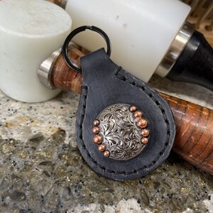 May include: A black leather keychain with a silver and copper concho featuring a floral design. The keychain has a silver key ring.