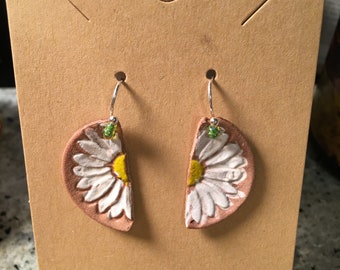 May include: A pair of handmade earrings featuring a half-circle design with a white daisy flower on a peach-coloured background. The earrings are made of leather and have silver hooks.