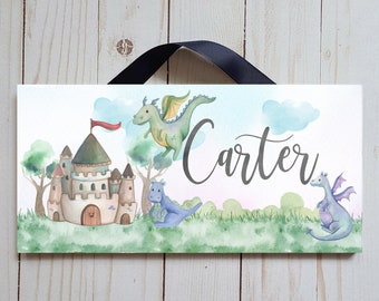 May include: A rectangular sign featuring a watercolor illustration of a castle, dragons, and the name "Carter". The sign has a black ribbon for hanging. The artwork includes green, blue, and pink hues, creating a whimsical, fantasy theme.