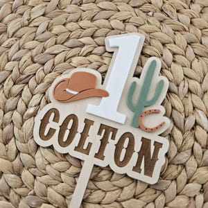 Peut inclure: Un cake topper en bois sur le th&egrave;me du cowboy. Il pr&eacute;sente le chiffre 1, un chapeau de cowboy, un cactus et le nom "COLTON". Le topper est dans les tons de marron, blanc et vert, parfait pour un premier anniversaire.