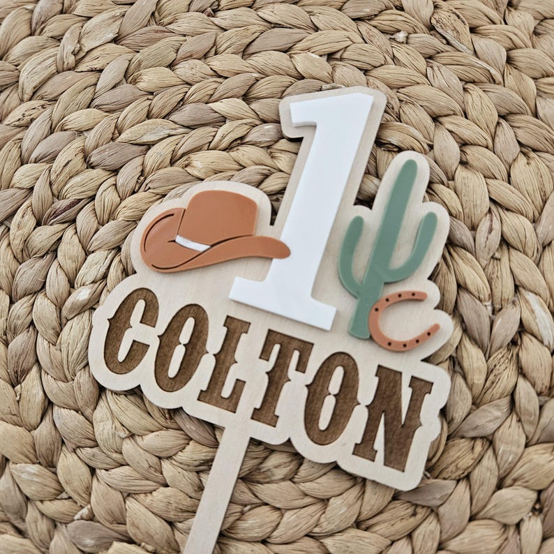 Peut inclure: Un cake topper en bois sur le th&egrave;me du cowboy. Il pr&eacute;sente le chiffre 1, un chapeau de cowboy, un cactus et le nom "COLTON". Le topper est dans les tons de marron, blanc et vert, parfait pour un premier anniversaire.