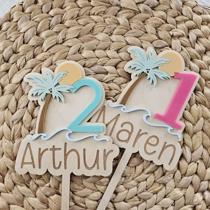 May include: Two wooden cake toppers featuring palm trees, suns, and ocean waves. One topper displays the number 2 in light blue and the name Arthur. The other topper displays the number 1 in pink and the name Maren, both with wooden stakes.