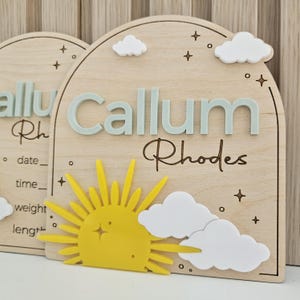 May include: An arched wooden plaque featuring the name Callum Rhodes in raised light blue lettering. The design includes a bright yellow sun, white acrylic clouds, and engraved star accents. The plaque also displays blank lines for date, time, weight, and length.