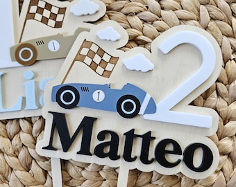 Pode incluir: Toppers de bolo de madeira com carros de corrida, bandeiras quadriculadas e nuvens. Um topper mostra o nome "Matteo" e o número "2". Os carros são azuis e bege. As bandeiras são castanhas e brancas.