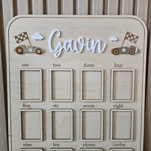 以下が含まれることがあります： 白い筆記体で「Gavin」という名前が入った木製の壁の装飾。ボードには、12個の長方形のフレーム、レーシングカー、雲、数字があります。レーシングカーには、市松模様の旗と数字の「1」が付いています。