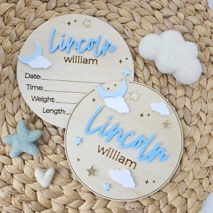 May include: Two circular wooden baby announcement plaques with the name "Lincoln William" in blue script. One has space for details like date and weight. Decorations include stars, moons, and clouds. Felt star, cloud, and heart also visible.