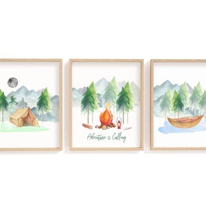 May include: Set of three framed watercolor prints featuring camping scenes. Each print depicts a different outdoor scene: a tent under a moon, a campfire, and a boat on a lake. The middle print has the text "Adventure is Calling".