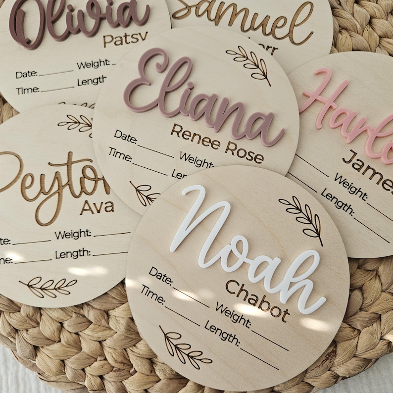May include: A collection of circular wooden birth announcement plaques featuring raised acrylic names in various colors. Each plaque includes engraved lines for date, time, weight, and length, accompanied by delicate leaf illustrations.