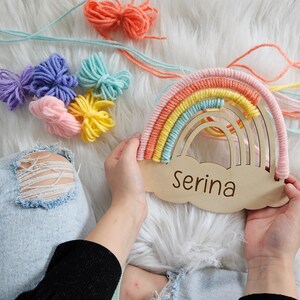 May include: A wooden rainbow-shaped wall hanging with a cloud base, personalized with the name "Serina." The rainbow features pastel colors of pink, yellow, orange, and blue. Colorful yarn bundles and strands are scattered around the rainbow.
