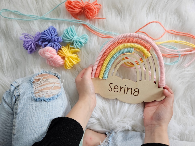 May include: A wooden rainbow-shaped wall hanging with a cloud base, personalized with the name "Serina." The rainbow features pastel colors of pink, yellow, orange, and blue. Colorful yarn bundles and strands are scattered around the rainbow.