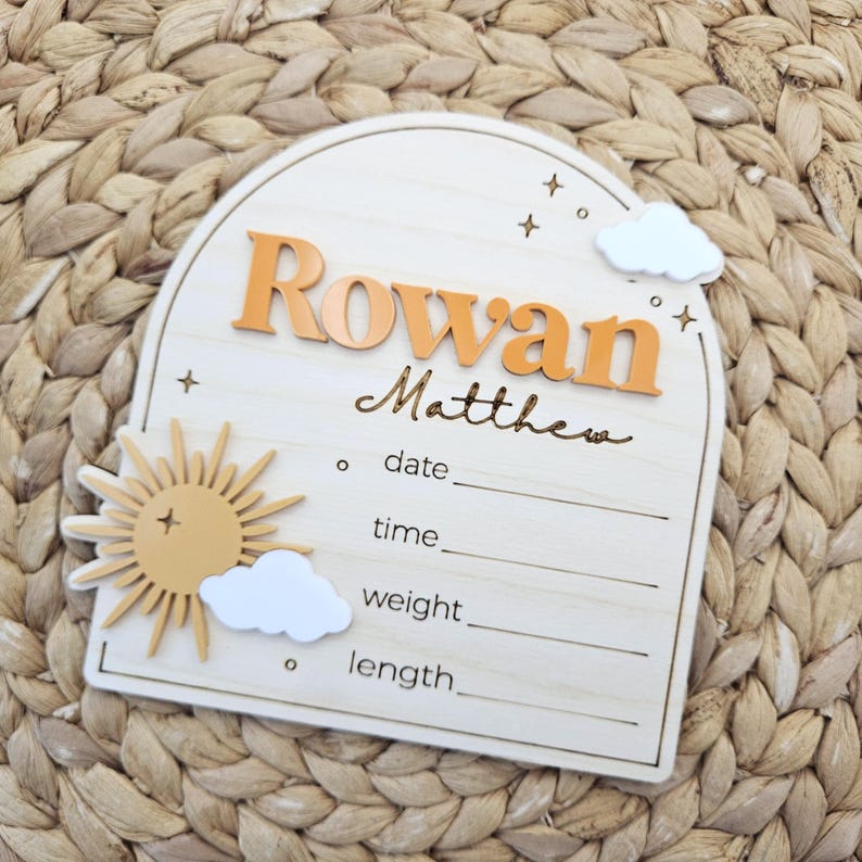 May include: An arched wooden birth announcement sign featuring the name Rowan Matthew in raised orange and script lettering. The plaque includes a yellow sun, white clouds, and engraved lines for date, time, weight, and length details.