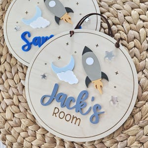 May include: Two circular wooden signs featuring space-themed decorations including a grey rocket, a light blue crescent moon, white clouds, and silver stars. One sign displays the blue text 'Jack's ROOM' and the other shows 'Sam'.