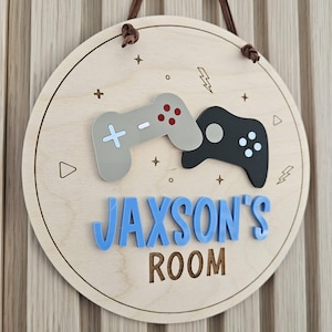 May include: A circular wooden wall sign featuring two layered gaming controller cutouts in light grey and black. The sign displays the blue acrylic text JAXSON'S and the engraved text ROOM, accented with small engraved lightning bolts and stars.