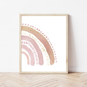 May include: A framed watercolor print featuring a pastel rainbow design. The rainbow arcs from the left, showcasing layers of pink, peach, and gold hues. The artwork is set against a white background, framed in a light wood.