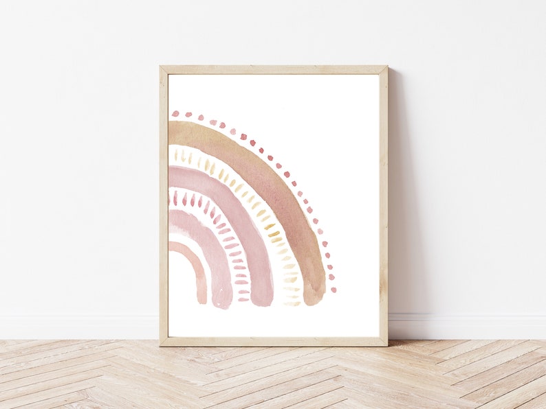 May include: A framed watercolor print featuring a pastel rainbow design. The rainbow arcs from the left, showcasing layers of pink, peach, and gold hues. The artwork is set against a white background, framed in a light wood.