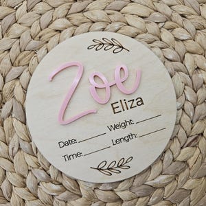 May include: A circular wooden plaque featuring the name Zoe in raised pink script lettering and the name Eliza in black text. Below, the words Date, Time, Weight, and Length are printed with blank lines for entry, framed by two etched leaf designs.