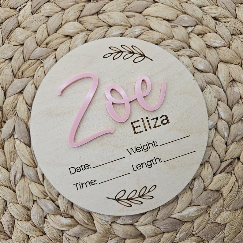 May include: A circular wooden plaque featuring the name Zoe in raised pink script lettering and the name Eliza in black text. Below, the words Date, Time, Weight, and Length are printed with blank lines for entry, framed by two etched leaf designs.