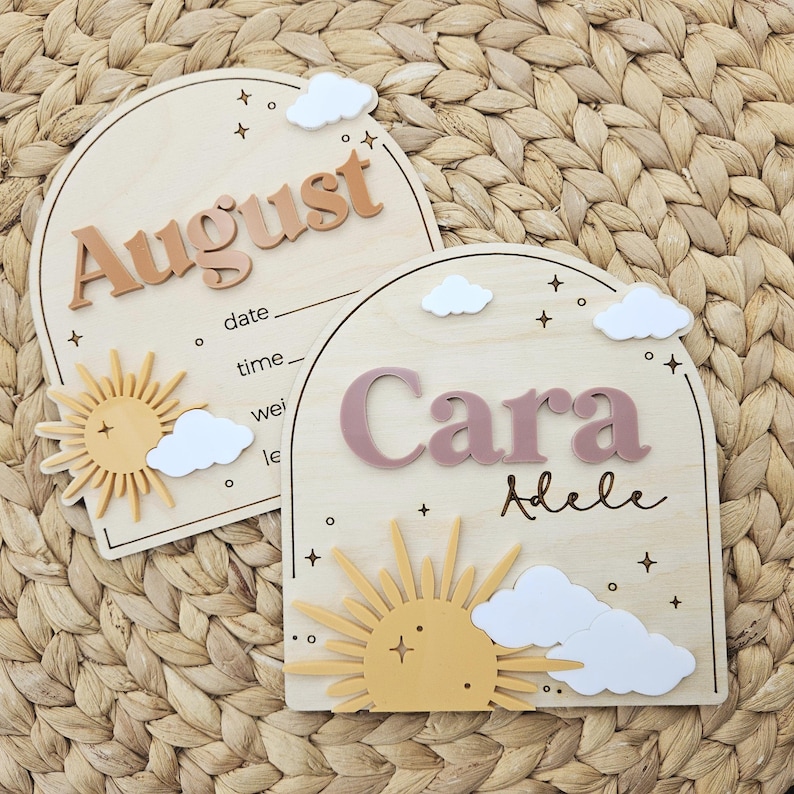 May include: Two arched wooden plaques featuring raised 3D lettering and sun and cloud motifs. One plaque displays the name August with printed lines for date, time, and weight. The other displays the name Cara Adele with a yellow sun and white clouds.