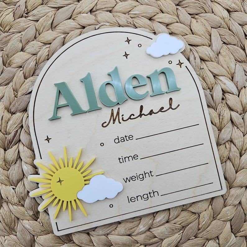 May include: An arched wooden birth announcement sign featuring the name Alden in raised sage green letters and Michael in brown script. It includes a yellow sun, white clouds, and engraved lines for date, time, weight, and length details.