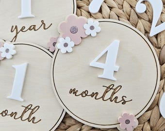 May include: Wooden milestone discs with white numbers and floral accents. The discs are light brown with laser-cut text, including "year" and "months." Pink and white flower embellishments surround the numbers. The background is a woven texture.