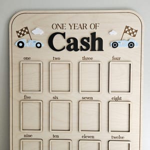 以下が含まれることがあります： 「ONE YEAR OF Cash」の文字が入った木製のマイルストーンボード。12個の長方形の写真スロット、レーシングカー、チェッカーフラッグ、雲が特徴です。1から12までの数字が写真スロットの下に印刷されています。