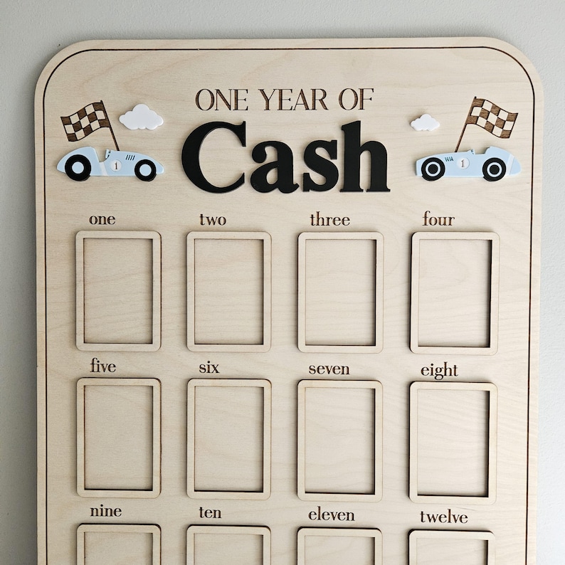 以下が含まれることがあります： 「ONE YEAR OF Cash」の文字が入った木製のマイルストーンボード。12個の長方形の写真スロット、レーシングカー、チェッカーフラッグ、雲が特徴です。1から12までの数字が写真スロットの下に印刷されています。